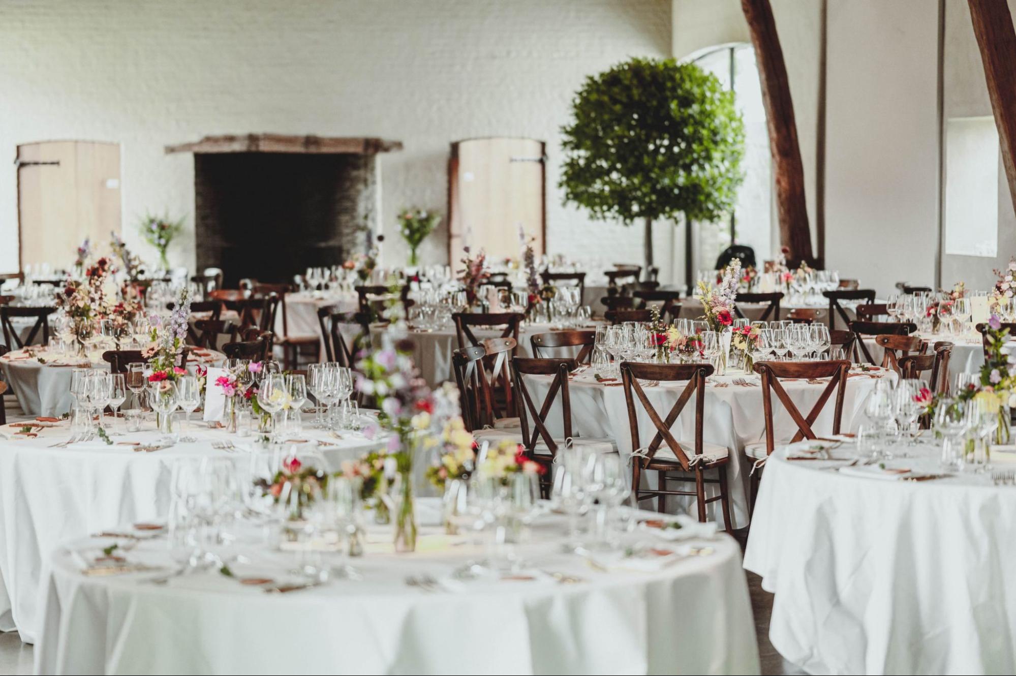 Feestzaal met ronde tafels, bloemen en gedekte couverts voor een huwelijksdiner
